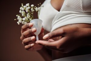 Image of a person, with a dark-toned skin, is holding a white colored menstrual cup. Inside the cup are small white flowers. The person is wearing a white colored top. 