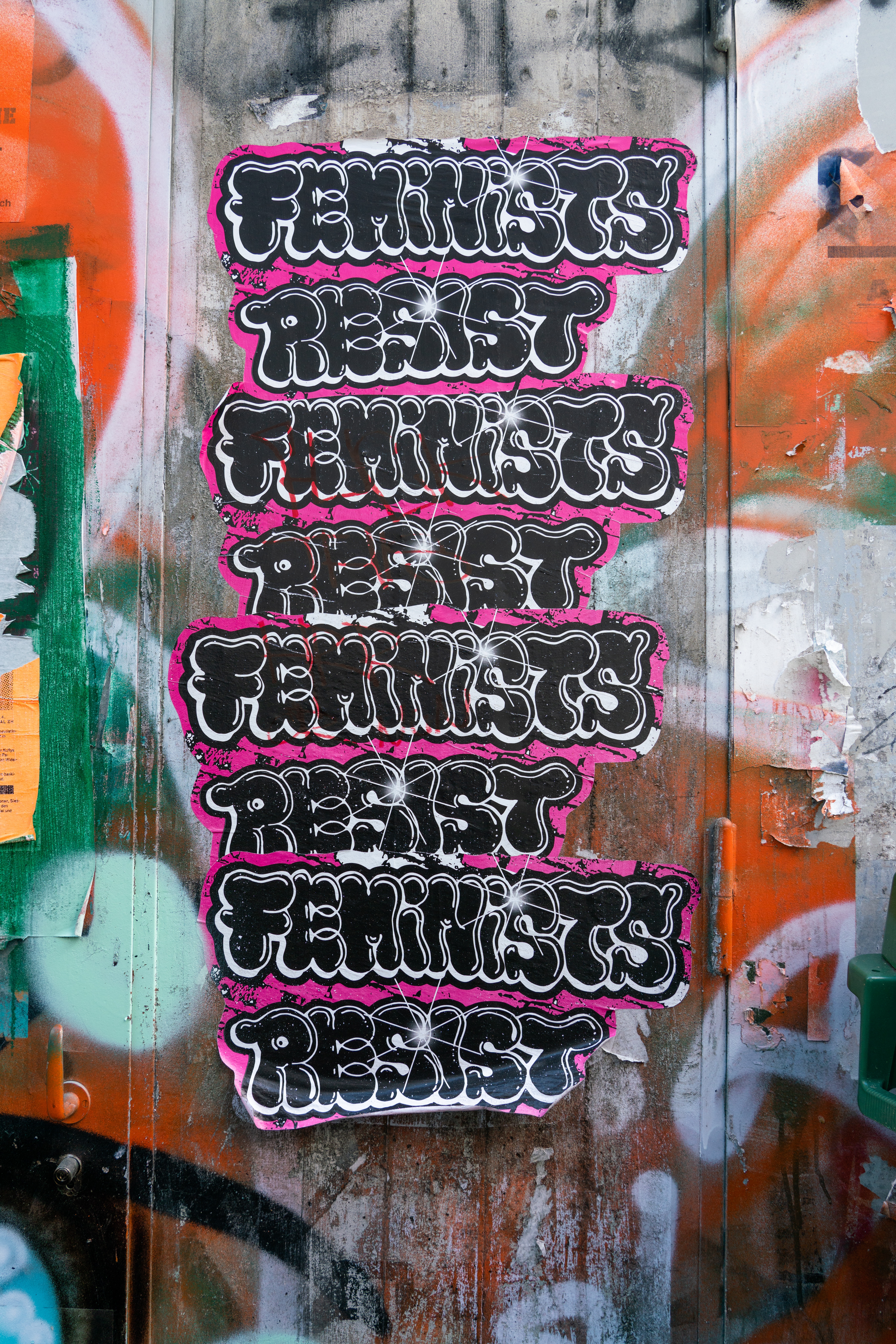 grafitti saying "feminist resist" repeatedly in black and pink color in a wall that is colorful 