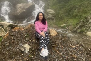 image of susmita, wearing a pink sweater with jeans. She is sitting on a rock with nature in the background